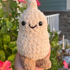Op de afbeelding: Een handgemaakt, pluche, beige gekleurd wezen met een lachend gezicht en kleine, bruine voetjes. Het knuffeldier heeft zwarte knoopogen en een gestikte mond. Het pluche speelgoed is ontworpen om op een teentje knoflook te lijken.