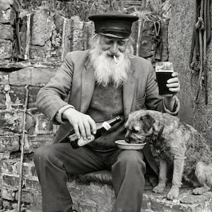 A vintage style photography print of a bearded man in a sailor cap sharing a beer with his terrier dog against a rustic stone wall. He holds a pint of stout while pouring a bottle into the dog's bowl.