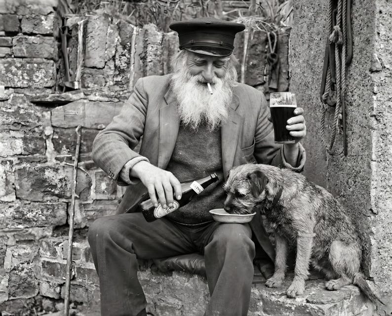 A vintage style photography print of a bearded man in a sailor cap sharing a beer with his terrier dog against a rustic stone wall. He holds a pint of stout while pouring a bottle into the dog's bowl.