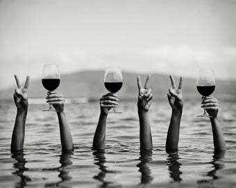 May include: Black and white image of arms raised from water, holding wine glasses filled with red wine. Some arms display peace signs. The scene is set against a blurred background of water and a distant shoreline.
