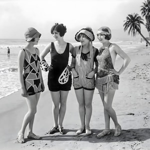 May include: Vintage black and white photograph of four women in swimwear standing on a beach. The women are wearing swimsuits, hats, and sandals. Palm trees and the ocean are visible in the background.