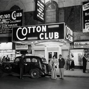 May include: Black and white photograph of the Cotton Club, a famous nightclub. The marquee reads "Cotton Club" with neon lights. A vintage car and people in formal attire are in front of the club. Signs advertise shows and dining.