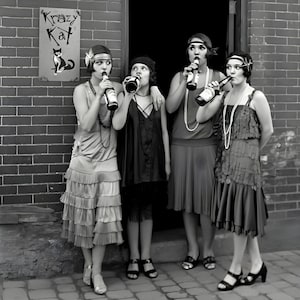 May include: Black and white image of four women in flapper dresses, each holding a bottle to their lips. They stand in front of a brick wall with a sign that reads "Krazy Kat" and a cat illustration. The women wear headbands and necklaces.