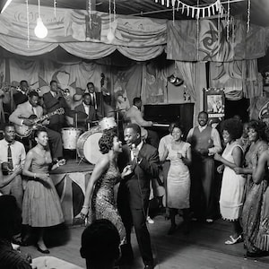 May include: Black and white photograph of a jazz band performing on a stage in a dimly lit club. People are dancing and enjoying the music. The band includes a drummer, guitar player, and a singer with a microphone.