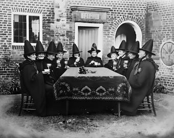 May include: A black and white photograph depicts a group of women seated around a table. They are dressed in dark robes and tall, pointed hats, reminiscent of witches. They are gathered outdoors, with a brick building in the background, and are holding glasses and cups.