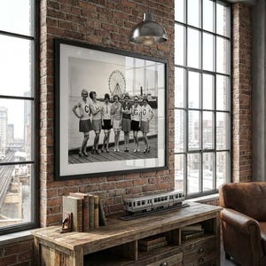 May include: Black and white framed photograph of a group of women in athletic attire posing on a pier. The photo is displayed on a brick wall, with a wooden console table below. A model train and books are on the table.