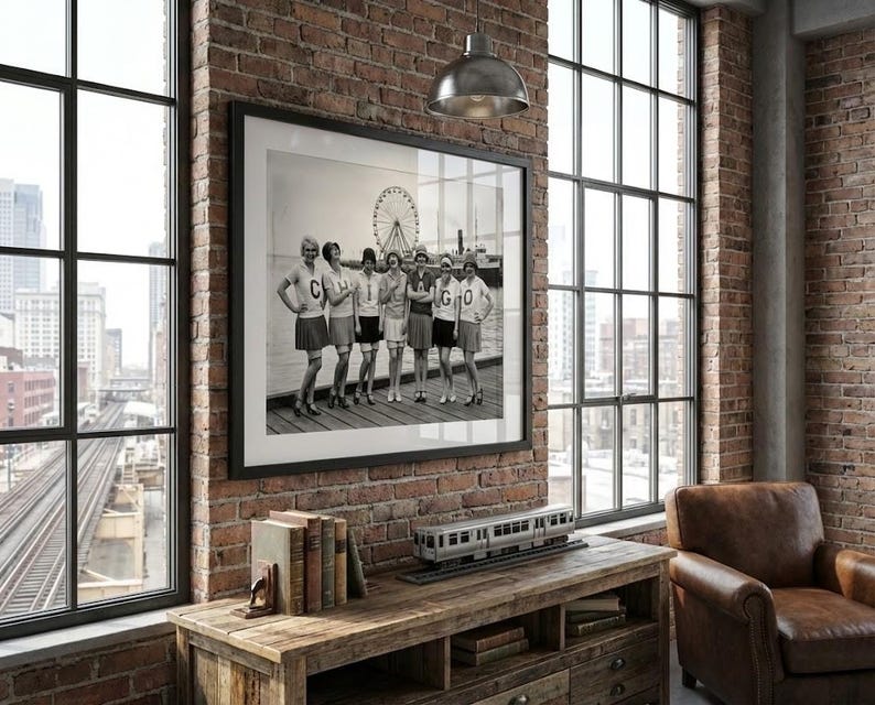 May include: Black and white framed photograph of a group of women in athletic attire posing on a pier. The photo is displayed on a brick wall, with a wooden console table below. A model train and books are on the table.