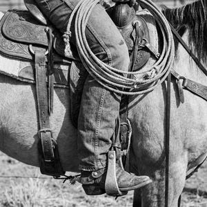 May include: Black and white image of a cowboy on horseback. The cowboy is wearing denim jeans and cowboy boots with spurs. A coiled rope rests on the saddle. The saddle is intricately tooled with a floral design.