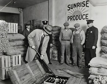 May include: Black and white photograph of a group of men in a warehouse. A man in a trench coat opens a wooden crate revealing bottles. Other men, including police officers, stand nearby. Sacks of produce and crates are visible.