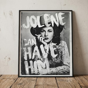 May include: Black and white framed print featuring a woman in a hat smoking a cigarette. The text "JOLENE CAN HAVE HIM" is painted in white over the image. The print is displayed on a wooden surface.