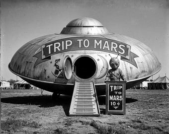 May include: A black and white photograph of a metallic, saucer-shaped attraction labeled TRIP TO MARS. A figure in an alien costume stands beside a sign reading TRIP TO MARS 10 cents. A ramp leads to an open circular hatch on the craft.