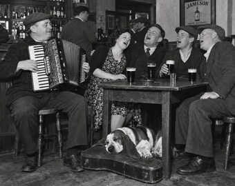 May include: A black and white photograph showing a group of people in a pub. One person plays a piano accordion while others sit at a wooden table with glasses of dark beer. A basset hound sleeps on a guitar case under the table. A Jameson sign is visible.