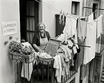 May include: A black and white photograph of a woman on a balcony doing laundry. Clothes hang on a clothesline, and a sign reads "LAVANDERIA DI MAMMA". A wooden washboard and a bowl of clothes are in front of her.