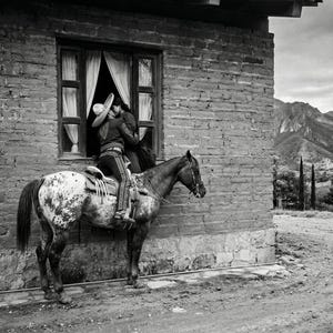 May include: Black and white photograph of a man in traditional charro attire on horseback, embracing a person in a window. The horse has a spotted coat. The scene is set against a brick building and a mountain backdrop.