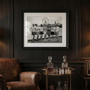May include: Black and white framed photograph of a group of people standing in front of a Ferris wheel. The photograph is hanging on a dark wood paneled wall. A leather armchair, a lamp, and a bar cart with decanters and glasses are in the foreground.