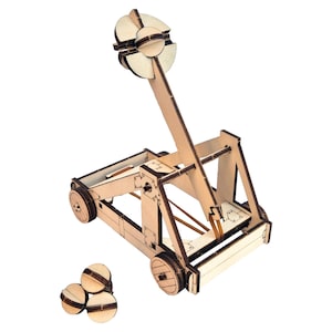 Könnte beinhalten: Ein helles Holzkatapult-Modell mit einem langen Wurfarm, einem kreisförmigen Projektilkorb und vier Rädern. Die Konstruktion besteht aus ineinandergreifenden Paneelen und einem Spannmechanismus. Drei kleine kreisförmige Holzprojektile liegen bei.