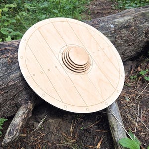 May include: A circular wooden shield featuring a light tan natural wood grain finish. The shield has a central raised boss composed of three stacked concentric circles and a perimeter border marked with small engraved circles along the outer edge.