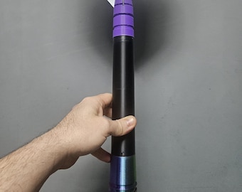 May include: A black and purple dart with a white and purple Snakebite Hardcore flight. The dart is held by a hand. The dart has a black barrel with a purple and iridescent grip. The flight has the text "Snakebite" and "Hardcore".