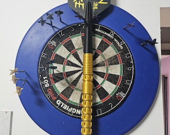 May include: A dartboard with a blue surround, featuring a black and gold dart in the center. The dart has a gold barrel and a black shaft, with a black and yellow "NUKE" flight. Several darts are embedded in the board.