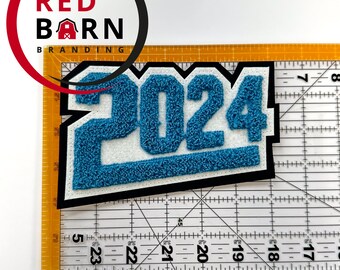 May include: A blue and white 2024 patch with a black border. The numbers are in a textured, chenille-like font. The patch is on a clear ruler with centimetre markings. The Red Barn Branding logo is in the upper left corner.