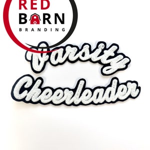 以下が含まれることがあります： 黒いアウトラインで書かれた、筆記体で「Varsity Cheerleader」と書かれた白いアイロン接着パッチです。