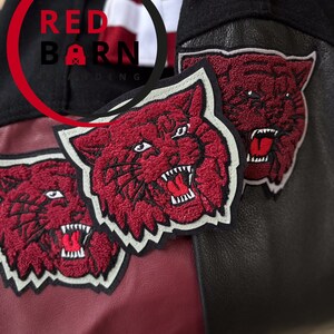 May include: Three red embroidered patches of a snarling tiger head on a black and burgundy leather jacket. The patches have white outlines and are sewn onto the jacket. The jacket has a red barn logo on the left side of the chest.