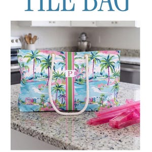 May include: A collection of mahjong accessories featuring a tropical palm tree print in blue, pink, and green. Items include a zippered pouch, a tote bag with white rope handles, and a square mat, all accented with a central monogrammed stripe.