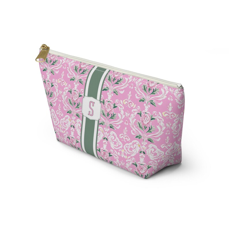 May include: A pink zippered pouch featuring a white and green damask floral pattern. A vertical sage green stripe runs down the center, containing a white rectangular label with the letter S printed in pink. The pouch has a gold-toned zipper.