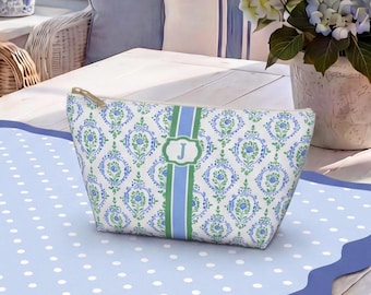 May include: A zippered pouch featuring a white background with a repeating blue and green floral damask pattern. A vertical blue and green striped band runs down the center, containing a rectangular frame with the letter J printed in white.