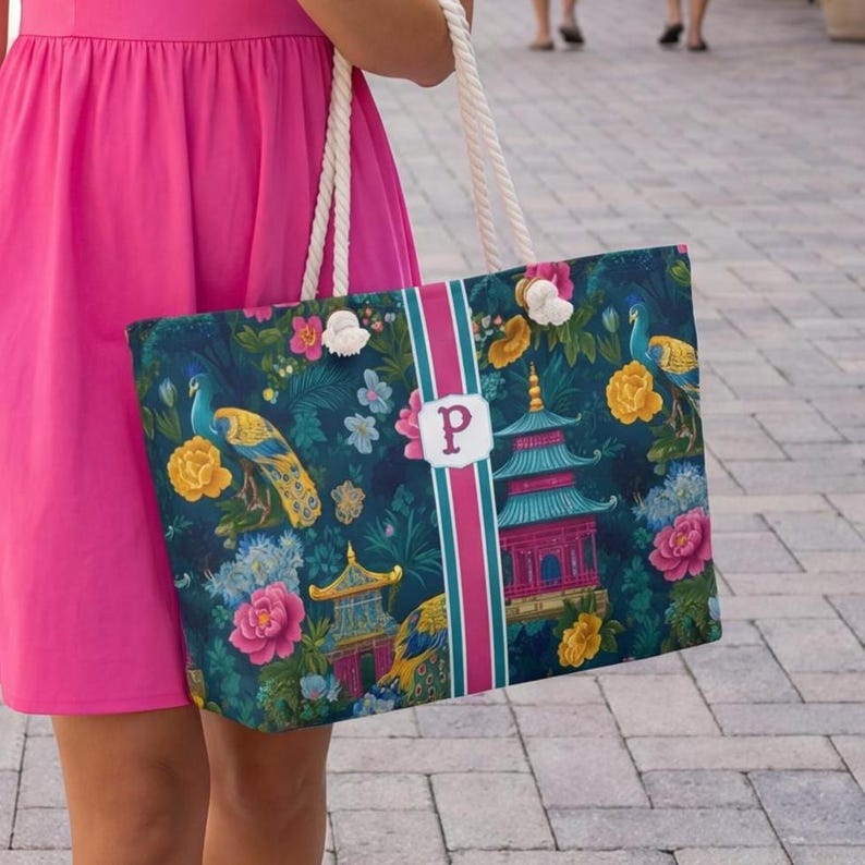 May include: A tote bag featuring a dark blue background with a pattern of colorful peacocks, yellow and pink flowers, and pagodas. A vertical stripe in pink and teal runs down the center with a white rectangular patch displaying the letter P.