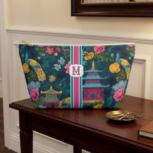 May include: A zippered pouch featuring a dark teal background with a pattern of colorful peacocks, pink and yellow flowers, and pagodas. A vertical pink and teal stripe runs down the center, displaying a white rectangular emblem with the letter M.