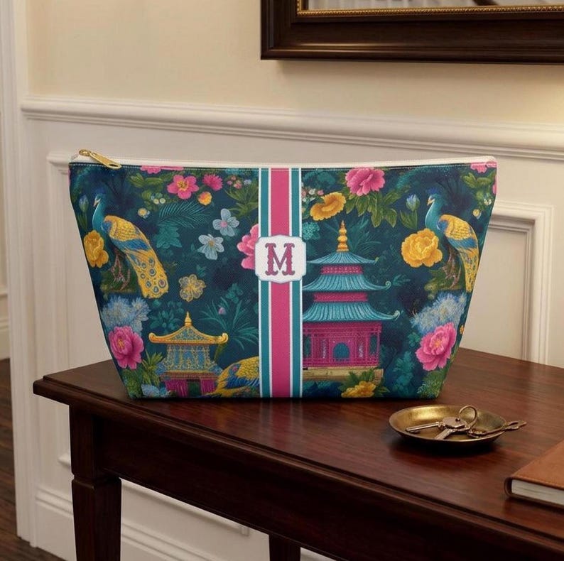 May include: A zippered pouch featuring a dark teal background with a pattern of colorful peacocks, pink and yellow flowers, and pagodas. A vertical pink and teal stripe runs down the center, displaying a white rectangular emblem with the letter M.