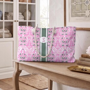 May include: A rectangular tote bag featuring a pink and white floral damask pattern with a central vertical dark green stripe. The bag includes white rope handles and a white rectangular emblem in the center displaying the letter S in pink.