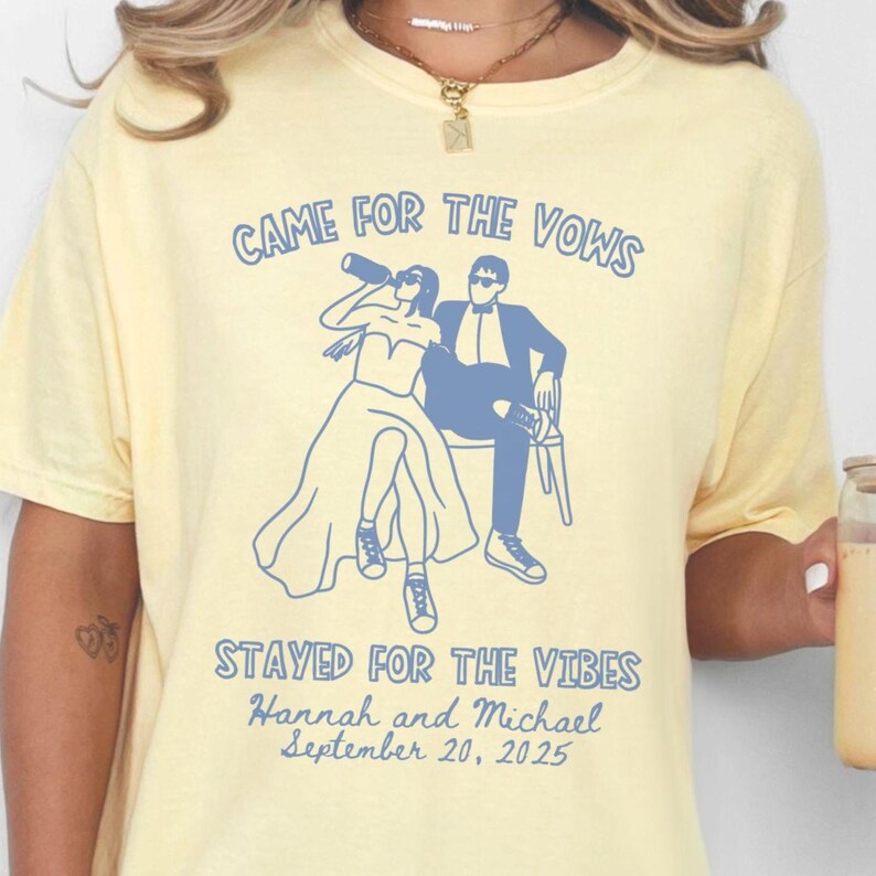 May include: A light yellow t-shirt with a blue graphic of a bride and groom, with the text "CAME FOR THE VOWS" above the graphic. Below the graphic is the text "STAYED FOR THE VIBES" and the names "Hannah and Michael" with the date "September 20, 2025".