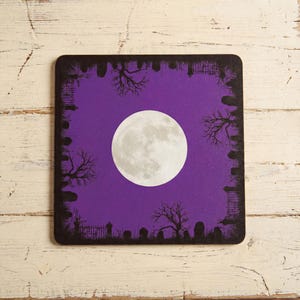 K&ouml;nnte beinhalten: Ein quadratischer Untersetzer mit einem leuchtend violetten Hintergrund und einem gro&szlig;en, zentrierten Vollmond. Der Rand ist mit schwarzen Silhouetten von kahlen B&auml;umen, Grabsteinen und Eisenz&auml;unen verziert, was eine dunkle, atmosph&auml;rische Szene schafft.