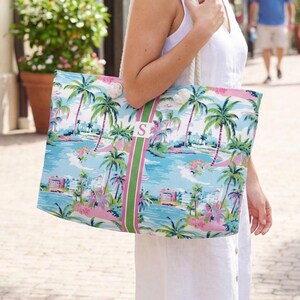 May include: A large tote bag featuring a tropical print with palm trees, pink buildings, and blue water. A vertical green and pink striped band runs down the center, displaying a white square patch with the letter S in a serif font. White rope handles.