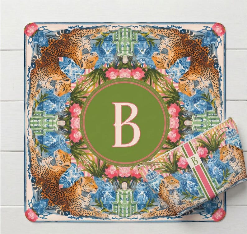 Puede incluir: Alfombra decorativa cuadrada con un c&iacute;rculo verde con la letra "B" en el centro. La alfombra tiene un dise&ntilde;o floral y de leopardo con detalles en azul y rosa. Tambi&eacute;n se ve una bolsa rectangular a juego.