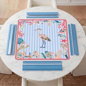 May include: A square play mat featuring a central heron illustration on a light blue and white striped background, surrounded by a border of colorful sea creatures including lobsters, crabs, and fish, framed by a pink edge and blue rails.
