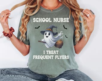 May include: A sage green t-shirt featuring a graphic of a white ghost wearing a witch hat and riding a broomstick. The text reads SCHOOL NURSE and I TREAT FREQUENT FLYERS, accompanied by small black bat illustrations. The shirt has a crew neckline.