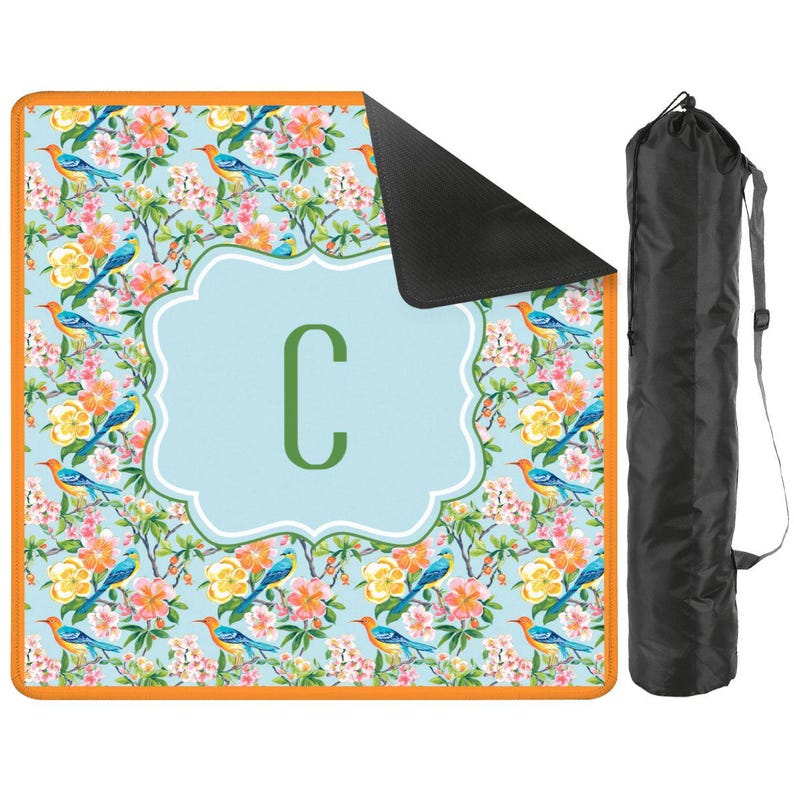 Puede incluir: Una manta de picnic con estampado floral con fondo azul claro, ribete naranja y reverso negro. La manta presenta una gran letra "C" en un marco azul claro. Se incluye una bolsa de transporte negra.