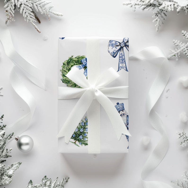 Puede incluir: Una caja de regalo rectangular envuelta en papel blanco con un patr&oacute;n azul y blanco de coronas, lazos, cascanueces y &aacute;rboles de Navidad. La caja est&aacute; atada con una cinta de sat&eacute;n blanca, ancha y lisa, dispuesta en un lazo central.