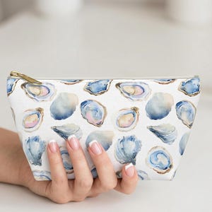 May include: White cosmetic bag with a watercolor oyster pattern in shades of blue, pink, and gold. The bag has a gold zipper and is held by a hand.