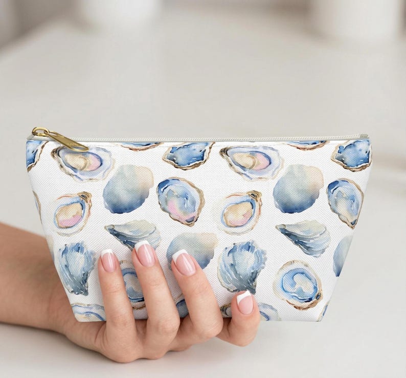 May include: White cosmetic bag with a watercolor oyster pattern in shades of blue, pink, and gold. The bag has a gold zipper and is held by a hand.