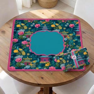 Op de afbeelding: Een vierkante bureaumat en een bijpassend rechthoekig etui met een donkergroenblauwe achtergrond, voorzien van een patroon met pauwen, roze pioenrozen, gele bloemen en pagodes. De mat heeft een effen blauwgroen middenvlak en een felroze rand.