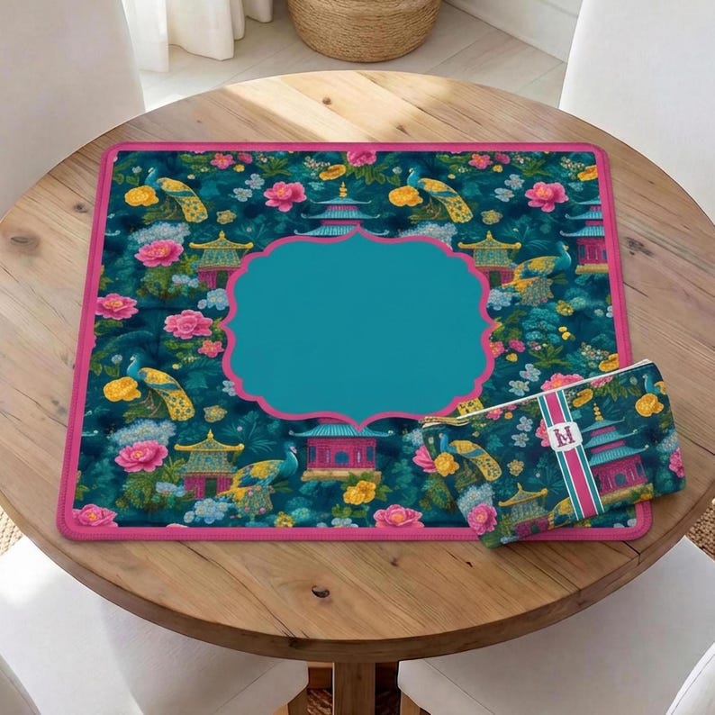 Op de afbeelding: Een vierkante bureaumat en een bijpassend rechthoekig etui met een donkergroenblauwe achtergrond, voorzien van een patroon met pauwen, roze pioenrozen, gele bloemen en pagodes. De mat heeft een effen blauwgroen middenvlak en een felroze rand.
