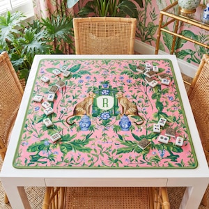 May include: A square white table with a pink floral patterned mat featuring two cheetahs, blue vases, and the letter "R". The table is surrounded by rattan chairs and a matching rattan chair back. Mahjong tiles are scattered on the mat.