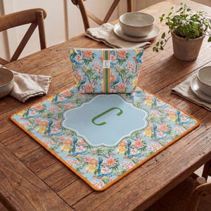 Puede incluir: Un conjunto de neceser y mantel individual con estampado floral. El fondo azul claro presenta p&aacute;jaros y flores. La bolsa y el mantel individual tienen una inicial de monograma. El mantel individual tiene un borde naranja.