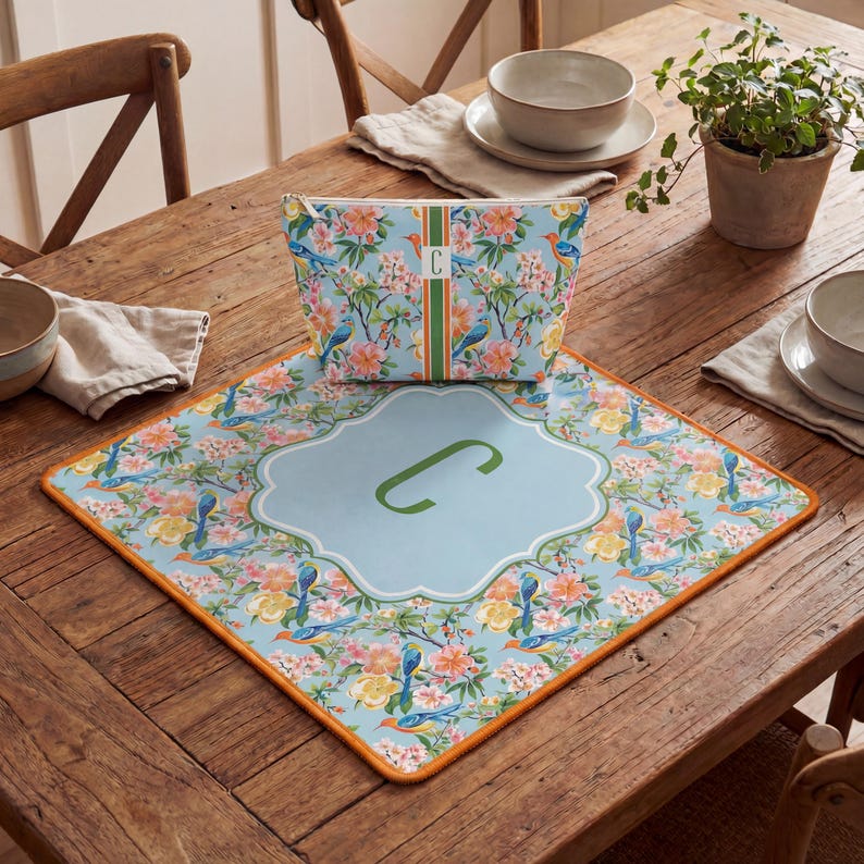 Puede incluir: Un conjunto de neceser y mantel individual con estampado floral. El fondo azul claro presenta p&aacute;jaros y flores. La bolsa y el mantel individual tienen una inicial de monograma. El mantel individual tiene un borde naranja.