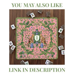 Puede incluir: Un tapete de Mahjong rosa con borde verde, con un dise&ntilde;o bot&aacute;nico con dos guepardos flanqueando un monograma. El tapete est&aacute; rodeado de fichas de Mahjong. El texto "YOU MAY ALSO LIKE" y "LINK IN DESCRIPTION" son visibles.