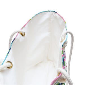May include: A tote bag featuring a vibrant tropical pattern with pink, green, and blue hues on the exterior. The bag has a solid white interior lining and thick, cream-colored rope handles attached with gold-toned metal grommets at the top.